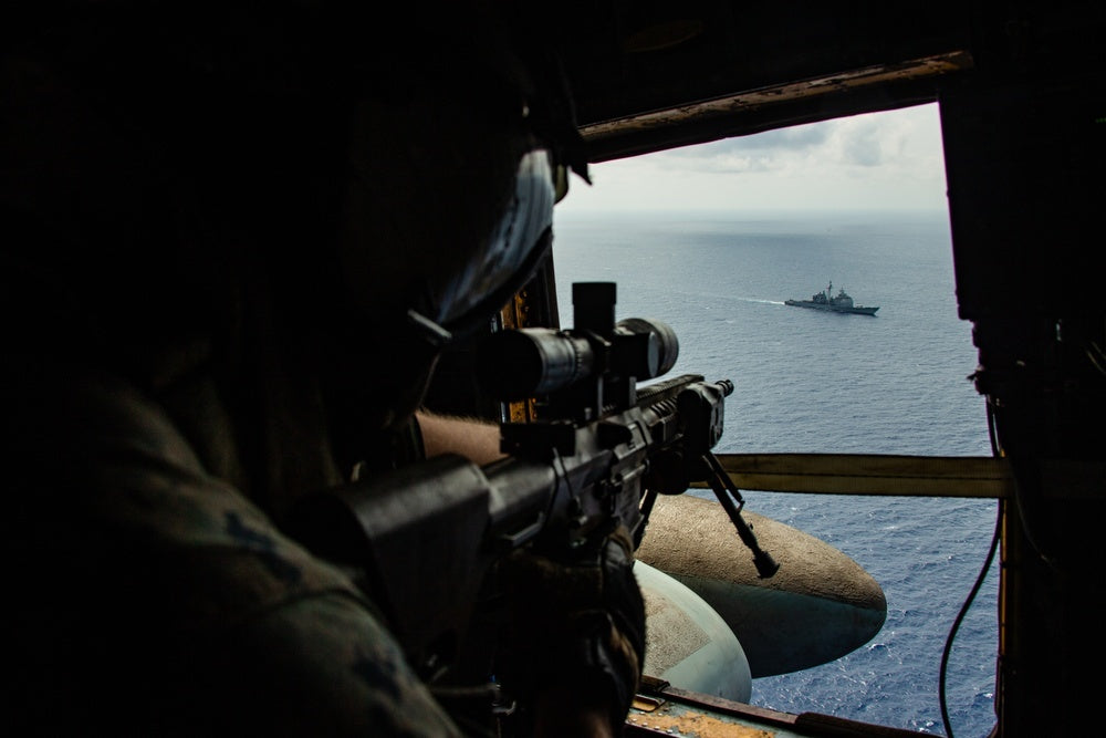 A U.S. Marine sights in on the Ticonderoga-class guided missile-cruiser USS Bunker Hill (CG 52) with an M110 sniper rifle out of the window of a CH-53E Super Stallion helicopter during a simulated visit, board, search and seizure (VBSS) drill.