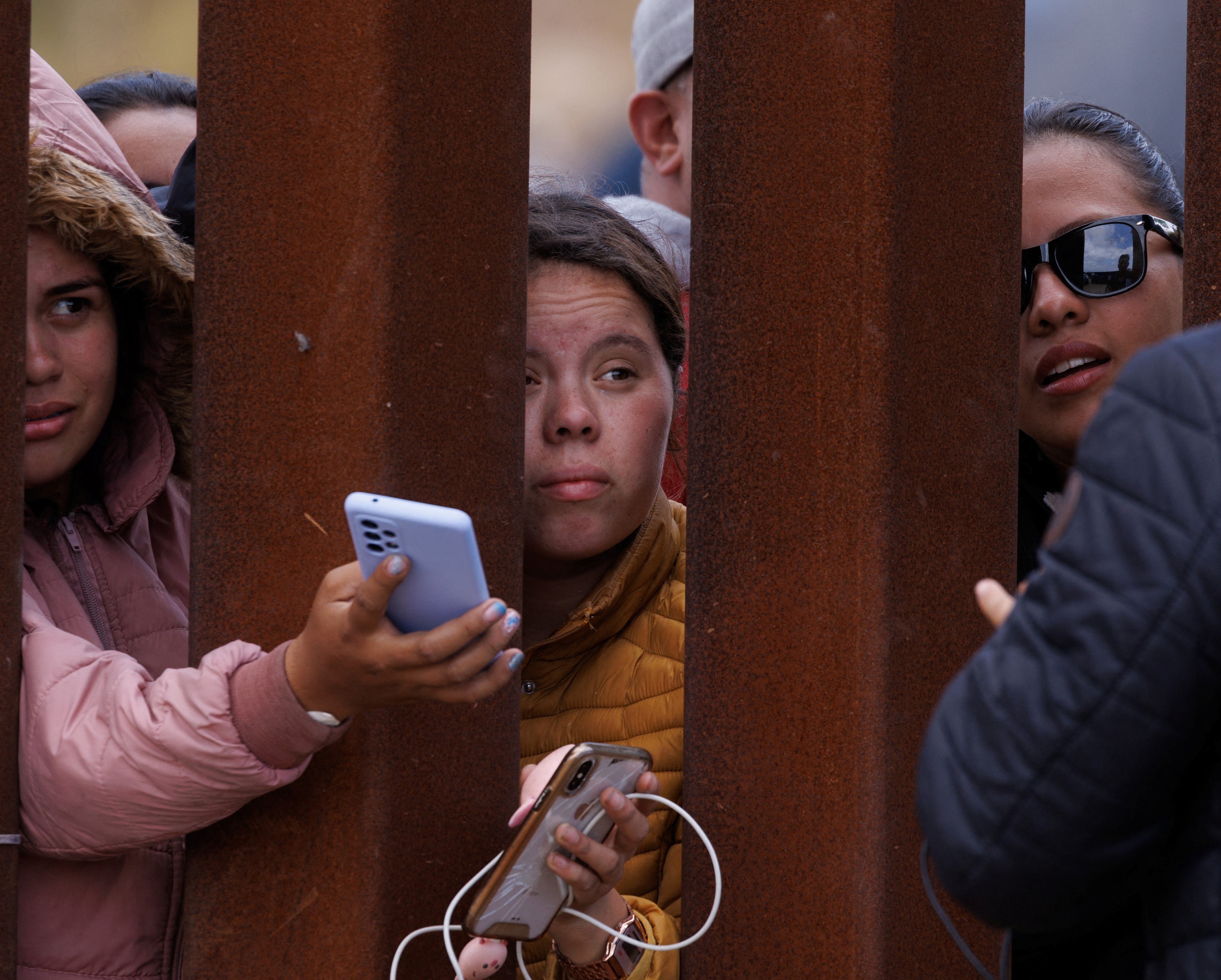 Migrants reach out from the border fence to try and get their phones charged while stuck between primary and secondary border fence